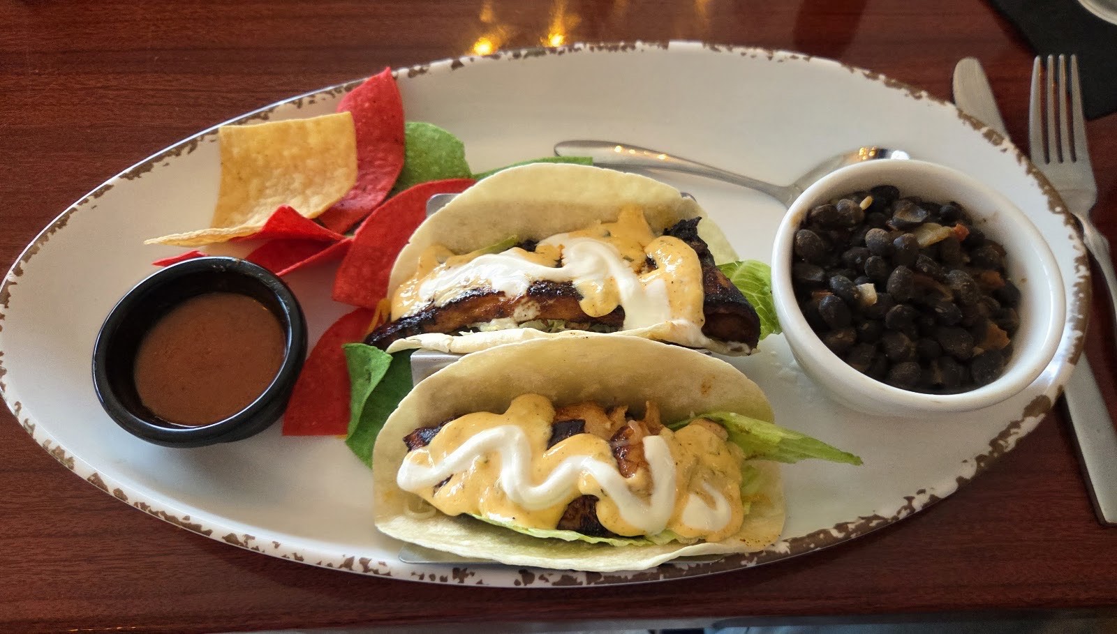 Two grilled fish tacos on soft tortillas with chipotle crema, crema drizzle, tortilla chips and a side of seasoned black beans